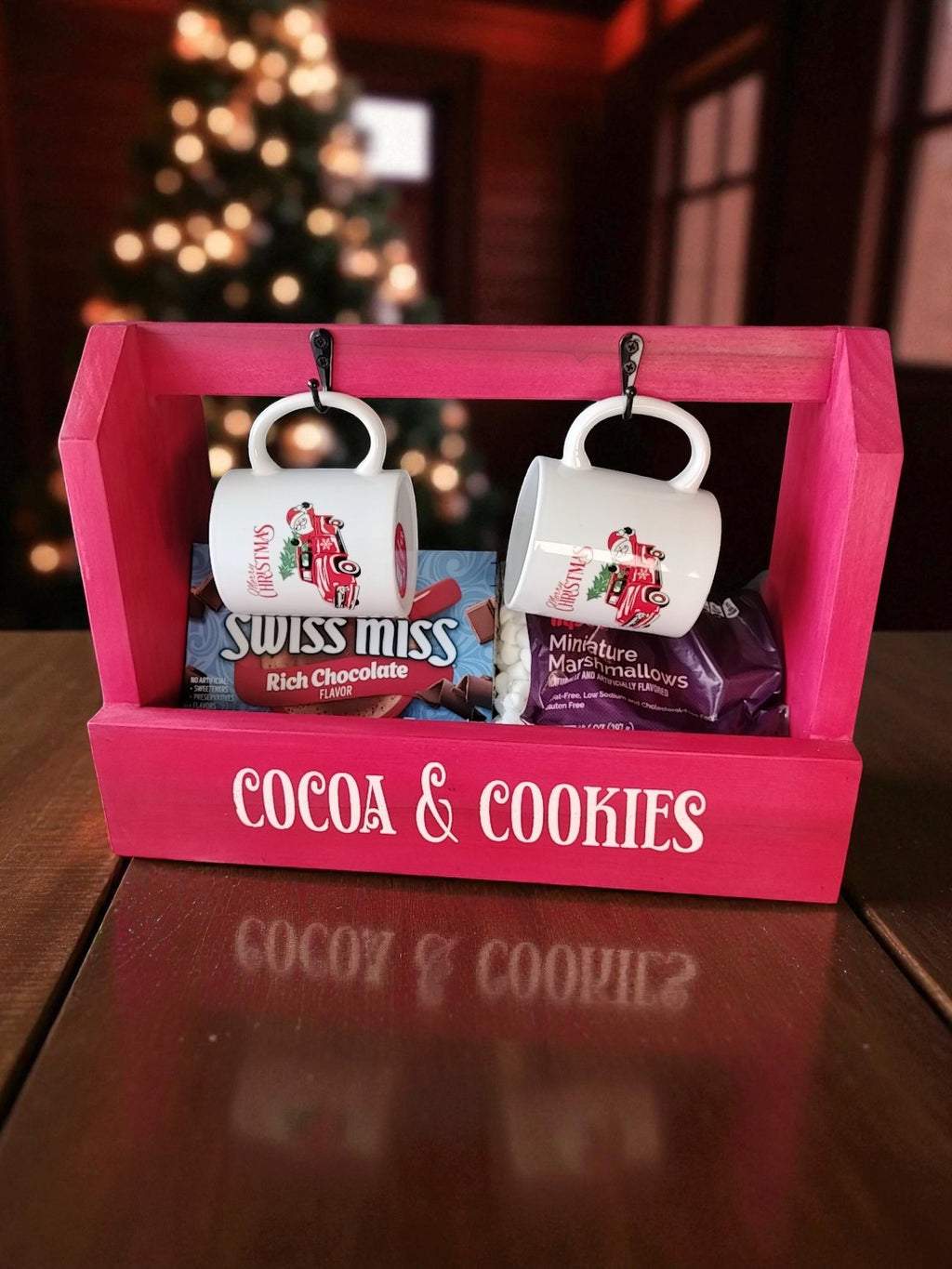 Cocoa & Cookies Caddy with two mugs cocoa mix and mini marshmallows contained in the caddy. Labeled 'Cocoa & Cookies', on a red wooden surface with a blurred Christmas tree in the background.