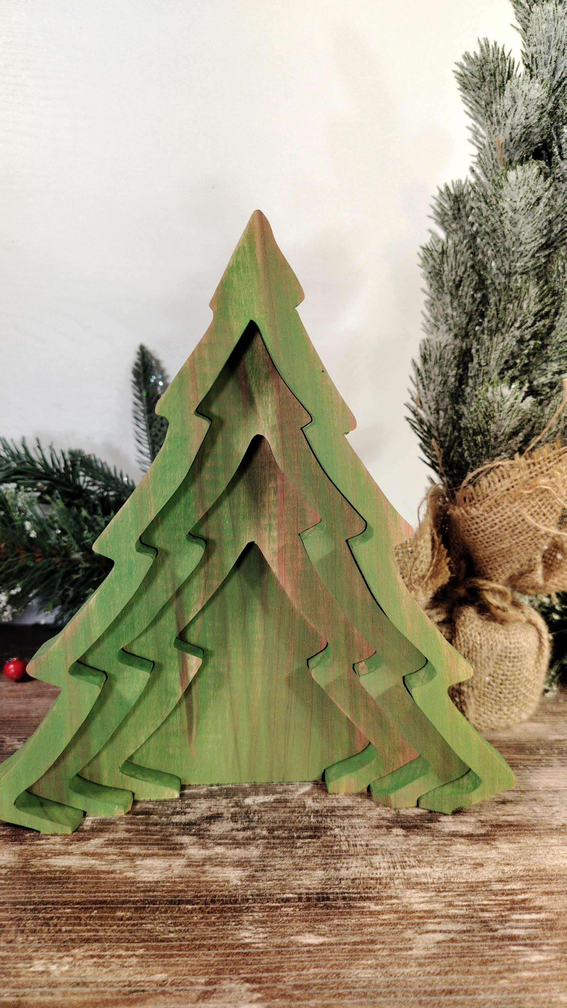 Green wooden Christmas tree decoration on a wooden surface with blurred Christmas trees in the background.
